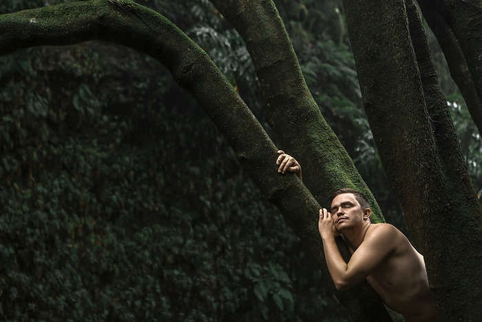 Shirtless man with eyes closed leaning on tree trunk