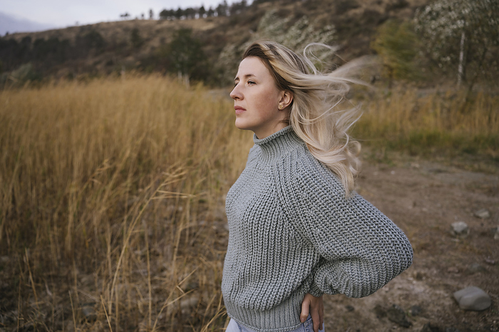 Blond woman wearing knitted sweater standing near dry tall grass