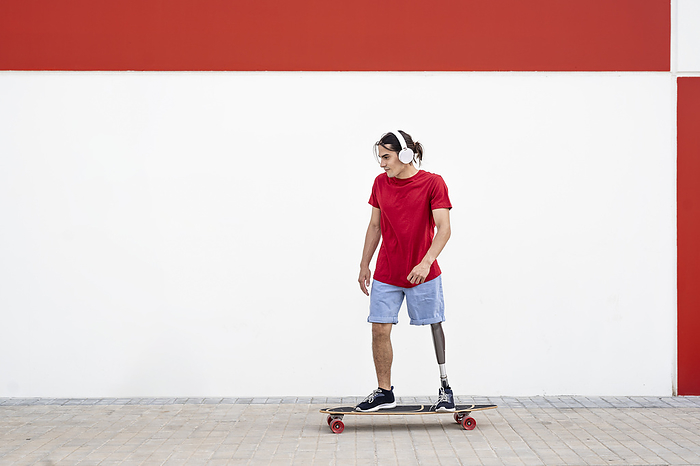 Young man wearing wireless headphones and skateboarding with prosthetic leg in front of wall