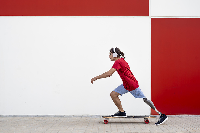 Man wearing wireless headphones and skateboarding with prosthetic leg in front of wall