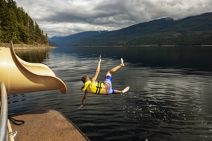 Young boy jumping out of the end of a waterslide on a houseboat on a fall day on Shuswap Lake; British Columbia, Canada, by LJM Photo / Design Pics