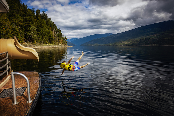Young boy jumping out of the end of a waterslide on a houseboat on a fall day on Shuswap Lake; British Columbia, Canada, by LJM Photo / Design Pics