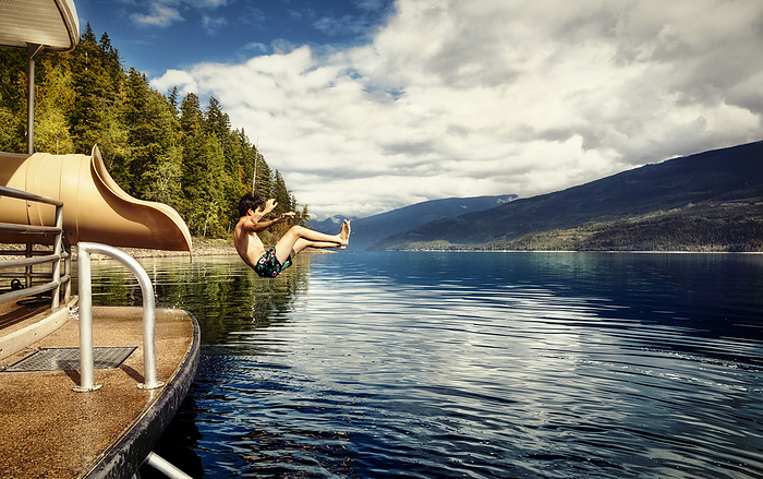 Boy jumping out of the end of a waterslide on a houseboat on a fall day on Shuswap Lake; British Columbia, Canada, by LJM Photo / Design Pics