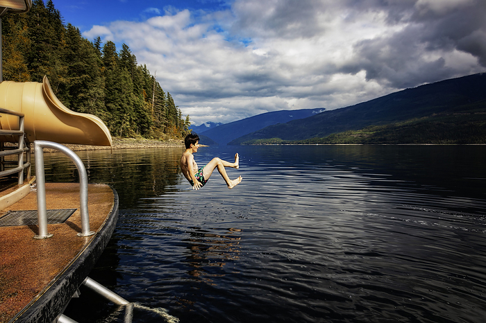 Boy jumping out of the end of a waterslide on a houseboat on a fall day on Shuswap Lake; British Columbia, Canada, by LJM Photo / Design Pics