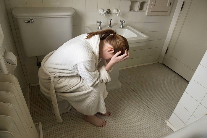 Young woman in a bathrobe sits uncomfortably in her bathroom; Lincoln, Nebraska, United States of America, by Joel Sartore Photography / Design Pics