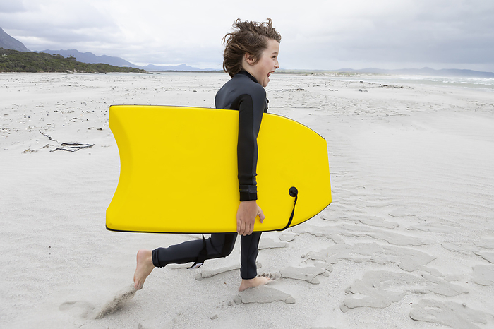 Boy (10-11) running on beach carrying body board, by Marc Romanelli
