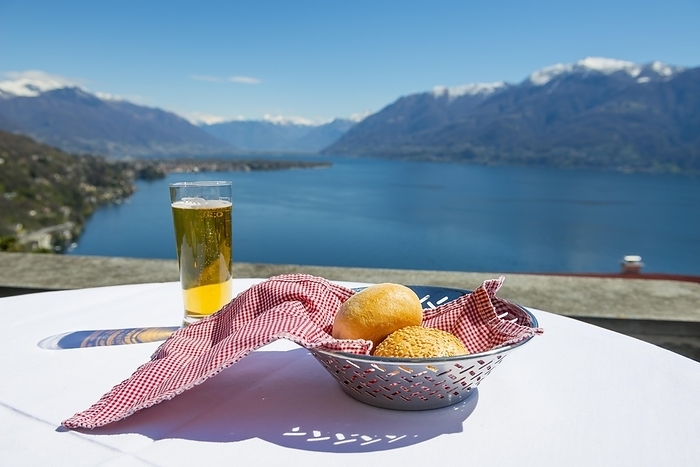 Table with Bread and a Glass of Beer With Panoramic View over Alpine Lake Maggiore with Snowcapped Mountain in a Sunny Day in Ticino, Switzerland, Europe, by Mats Silvan