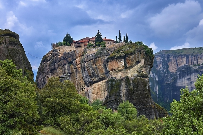 Close view of clifftop Eastern Orthodox monastery of Holy Trinity or Agia Triada in famous Meteora valley, Greece, listed as UNESCO World Heritage, among pinnacles of mountains, rich foliage, dramatic cloudy or stormy sky, Europe, by Natallia Pershaj