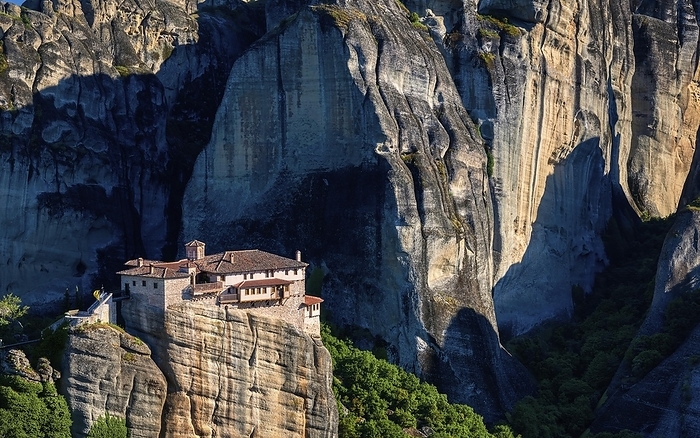 Close view of clifftop Moni Agias Varvaras Roussanou nunnery among hills covered with bushes and trees and massive sedimentary cliffs and rocks. Meteora valley, Greece at midday. UNESCO World Heritage, by Natallia Pershaj