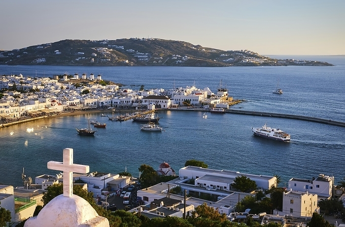 Beautiful sunset view over Chora, main town of Mykonos, Cyclades, Greece from roof top of a Greek Orthodox church, white marble cross. Golden hour, harbor, ships, white ferry arrive at port, whitewashed houses and distant islands in hazy background, by Natallia Pershaj