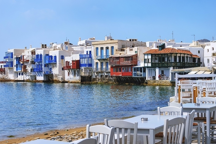 Little Venice of Mykonos main town or Chora, Cyclades, Greece. Romantic neighborhood with bars, cafes, restaurants in whitewashed old fisherman houses hanging on cliffs above sea waves. Colorful tables and chairs by shoreline. Summer day, blue sky, by Natallia Pershaj