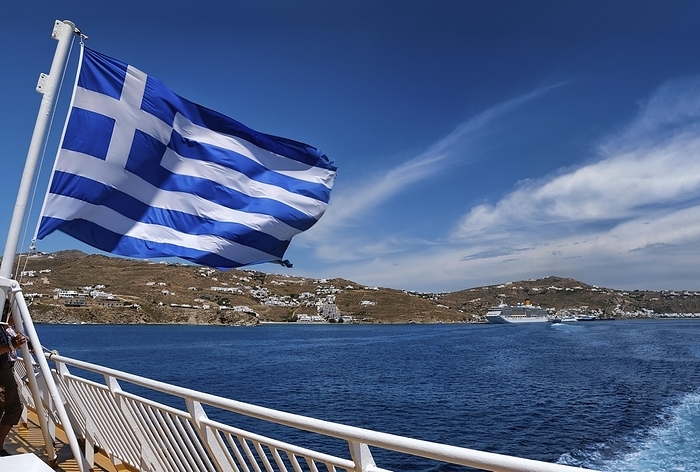 Greek flag waving against typical Greek island landscape. Cruise liners and ferries moored in background. Bright summer day. Island hopping, adventures and exploring Mediterranean sea. Selective focus, by Natallia Pershaj