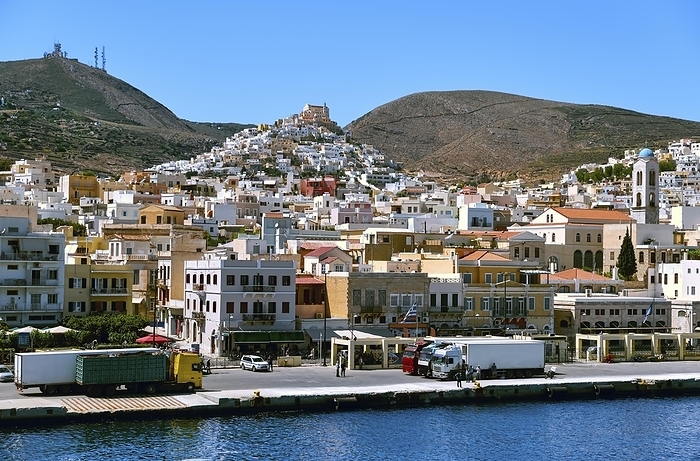 View of Ano Syros town, Syros island, Greece, and St George catholic cathedral on hilltop over medieval town on its slopes. Colorful houses, bright summer sunlight, trailer trucks wait for embarkation on ferry in port of Ermoupoli, Mediterranean sea, Europe, by Natallia Pershaj