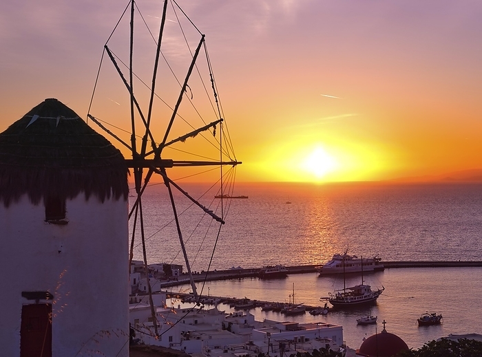 Famous traditional Greek windmill overlook port and harbor of Mykonos, Cyclades, Greece at sunset. Beautiful sunset sky, sun touch sea horizon, main town of island in sunset lights and shadows. Colorful image, selective focus, underexposed, by Natallia Pershaj