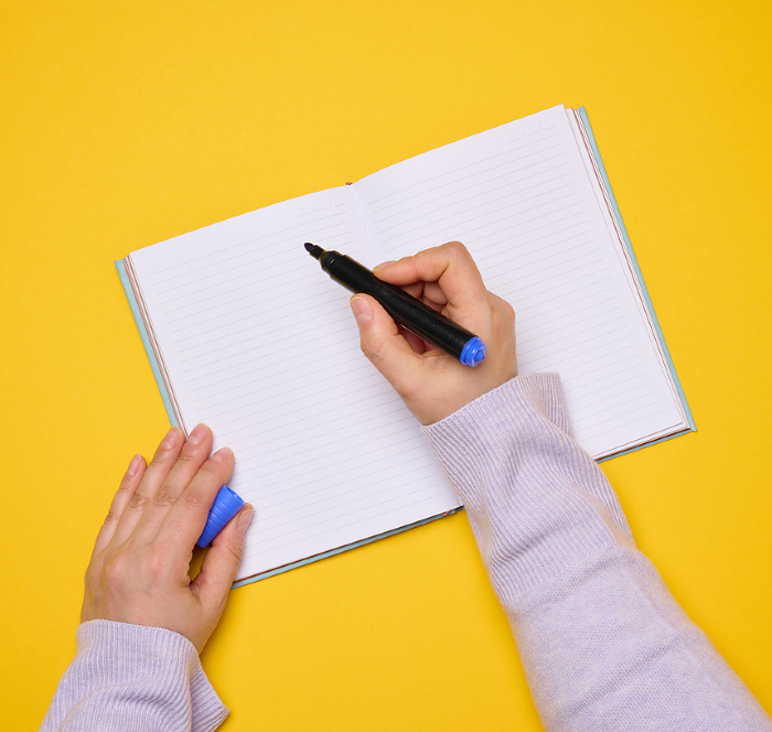 An open notebook with empty white sheets and a woman s hand holding a felt tip pen on a yellow background, top view An open notebook with empty white sheets and a woman s hand holding a felt tip pen on a yellow background, top view