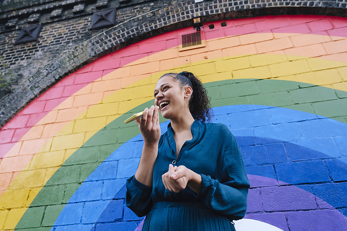 Happy young woman sending voicemail through smart phone in front of rainbow wall