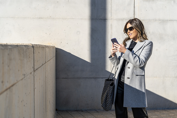 Smiling businesswoman wearing sunglasses and using smart phone in front of wall
