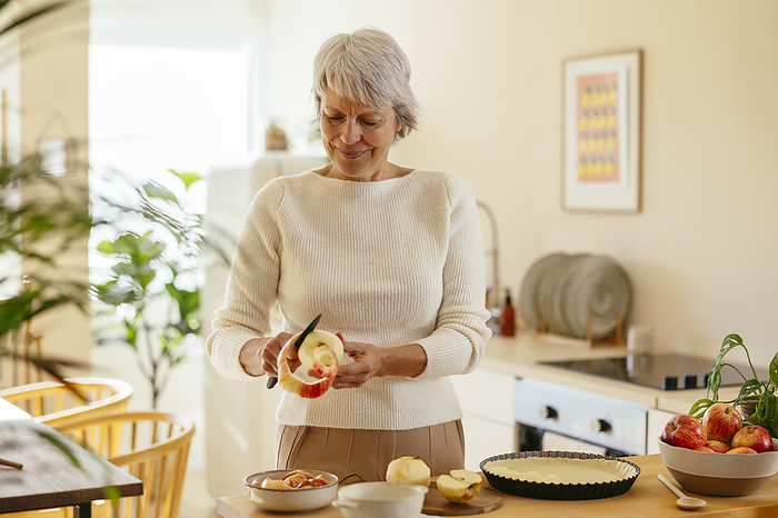 Mature woman peeling apples with knife at home