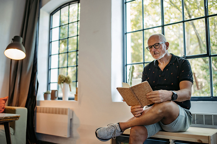Senior man in community space reading book sitting on bench