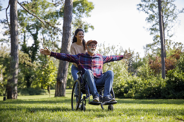Social worker pushing cheerful retired senior man with arms outstretched in wheelchair on grass at park