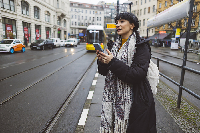 Smiling woman with smart phone standing at tram station