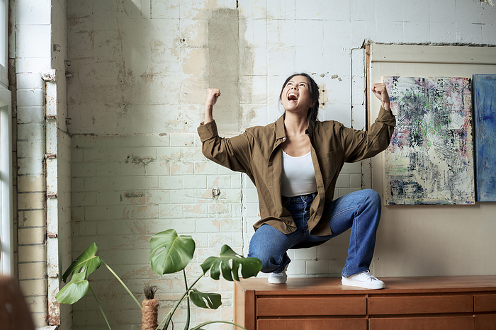 Excited painter shouting in joy squatting on cabinet by paintings at art studio