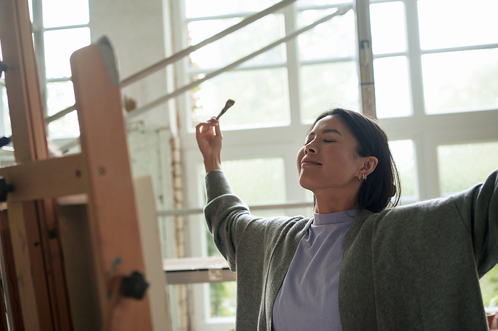 Smiling woman with eyes closed and arms outstretched standing in front of painting at workshop