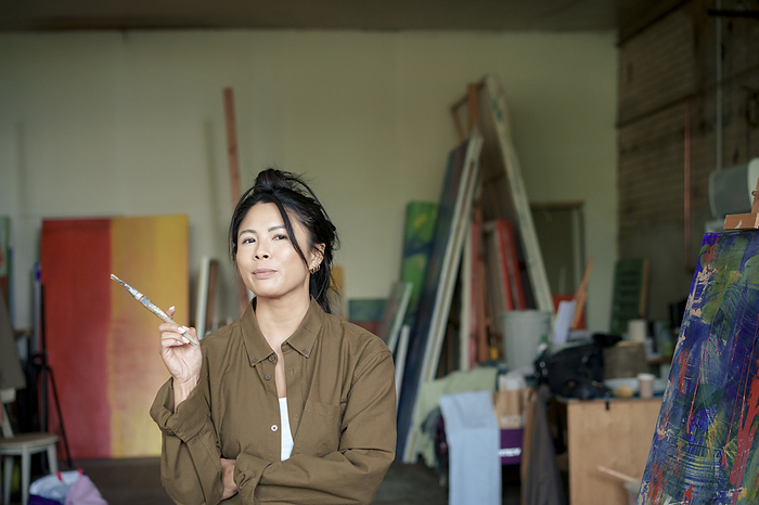 Confident painter holding paintbrush standing by painting at workshop