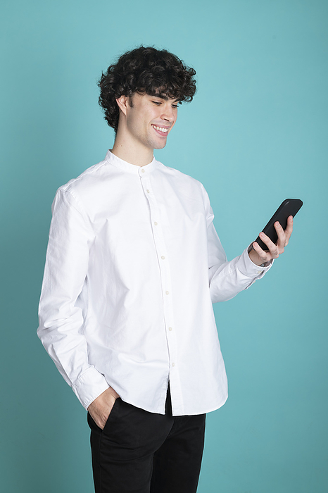 Smiling man with hand in pocket using smart phone standing against cyan background