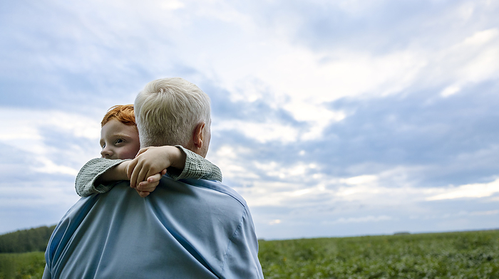 Grandfather carrying grandson in field under cloudy sky at sunset