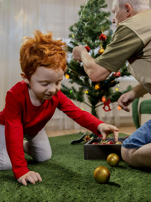 Redhead boy playing with bauble near grandfather decorating Christmas tree at home