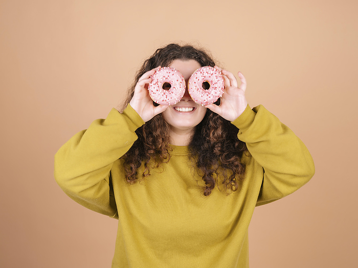Young woman playing with doughnuts Playful woman holding doughnuts over eyes against peach background