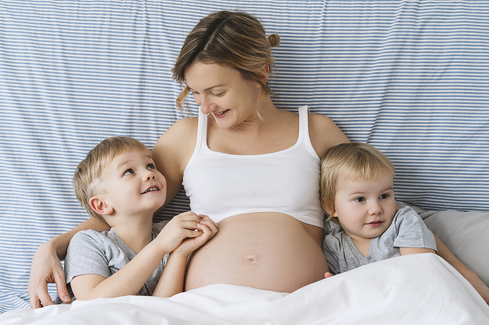 Smiling pregnant mother embracing children on bed at home