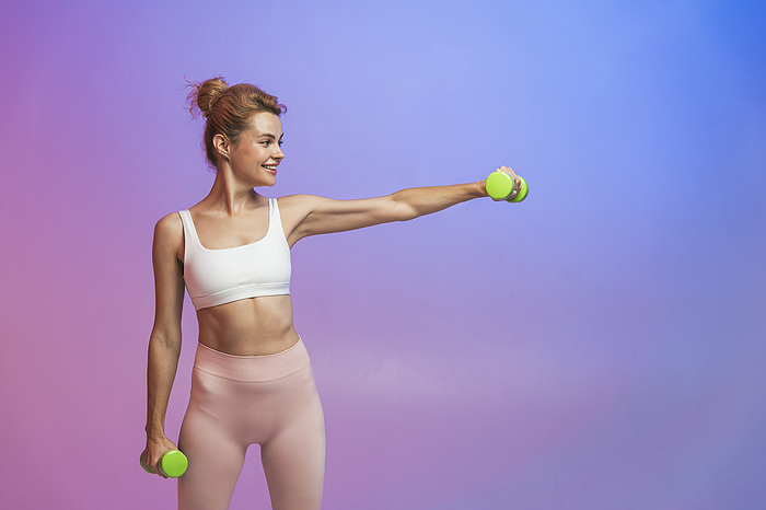 Smiling woman exercising with dumbbells standing against two tone background
