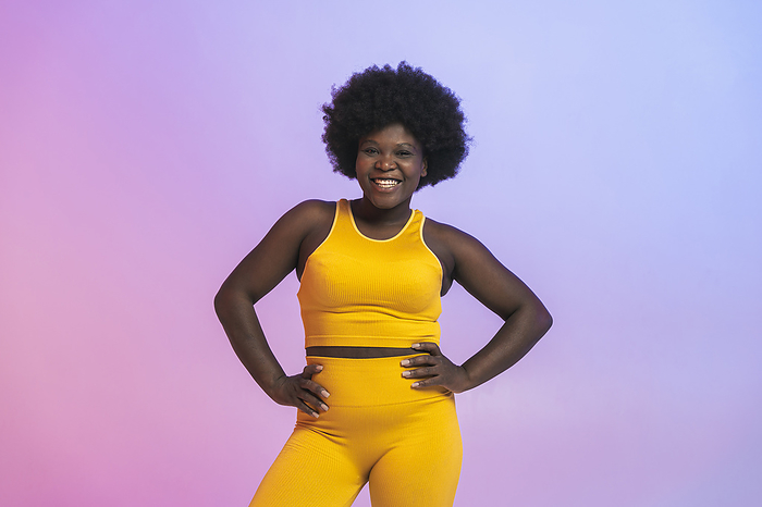 Happy young woman in yellow sports clothing standing with arms akimbo against two tone colored background