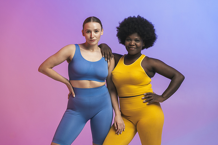 Confident young multiracial female friends standing with hands on hips against colored background