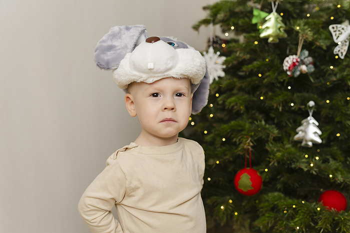 Sad boy standing near Christmas tree in front of wall at home