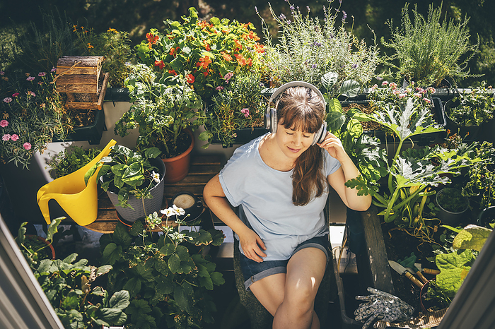 Smiling woman listening to music with wireless headphones near plants in balcony