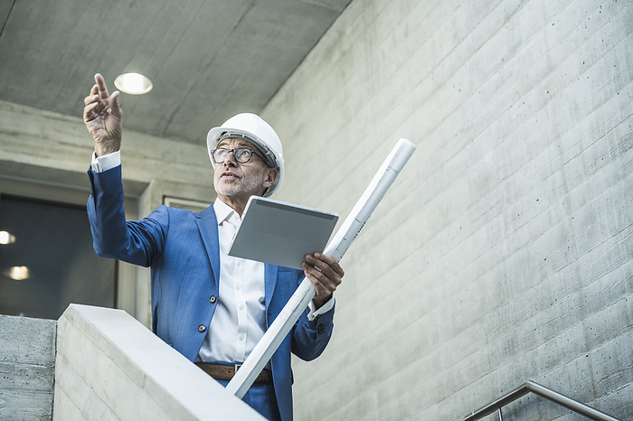 Engineer examining and holding tablet PC with blueprint near wall