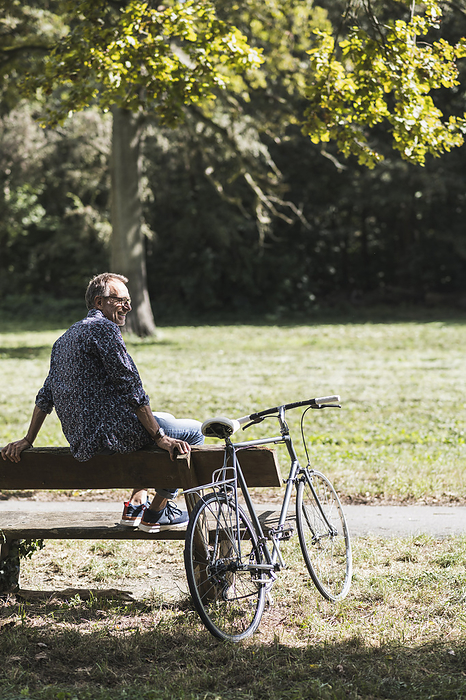 Senior man sitting on bench by bicycle in park on sunny day