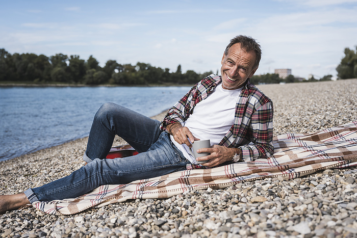 Smiling senior man with coffee cup sitting on blanket at beach