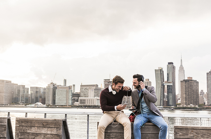 Happy business colleagues wearing headphones and listening to music in front of New York City skyline
