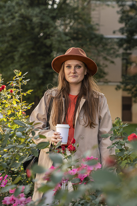 Smiling young woman wearing hat and standing with coffee cup near flowering plants