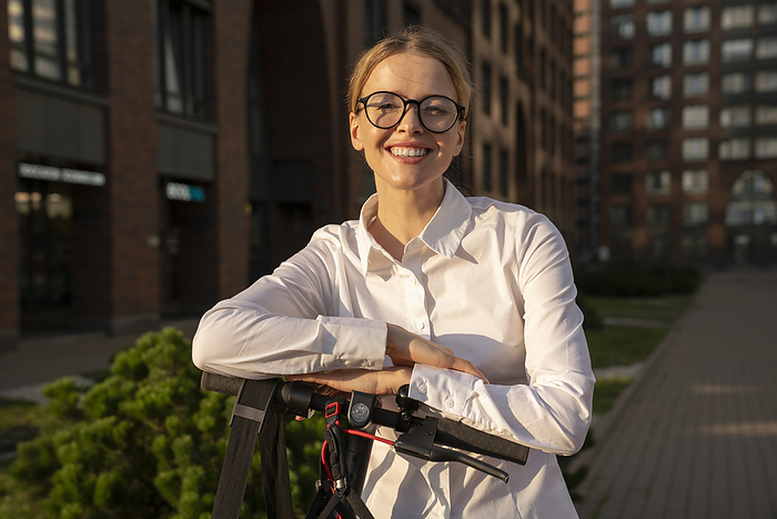 Happy businesswoman with electric push scooter and eyeglasses on footpath