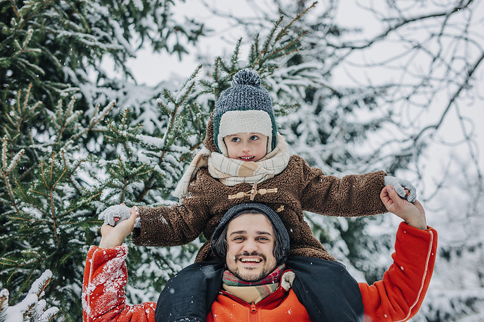 Happy man carrying son on shoulders and holding hands near tree