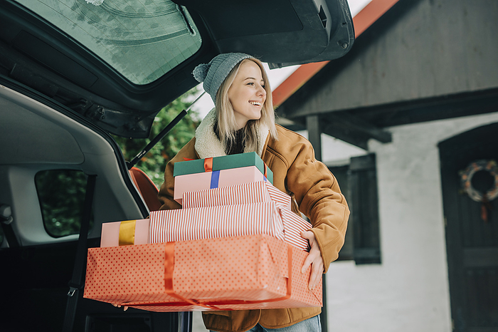 Smiling woman carrying Christmas presents near car trunk