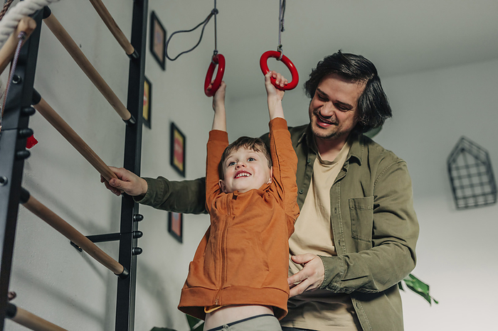 Smiling father teaching son to hang on gymnastic rings at home