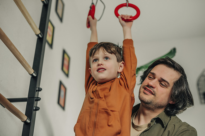 Happy father teaching son to hang on gymnastic rings at home