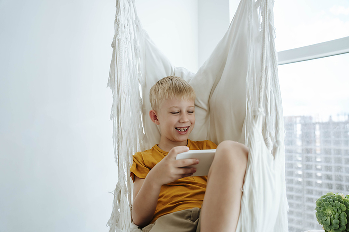 Happy boy sitting in hammock and using mobile phone at home