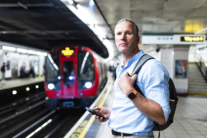 Mature businessman holding smart phone and waiting for train on platform